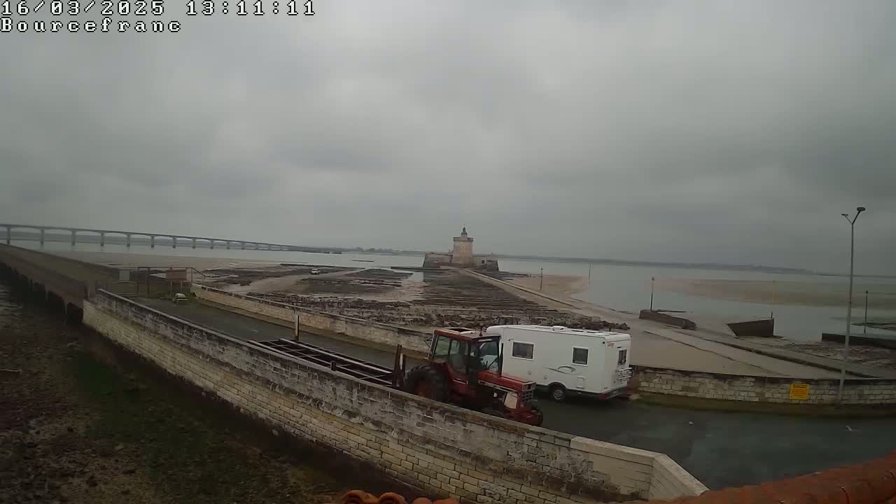 A red tractor and a white camper van are parked near a stone wall by the water, with a stone tower and a long bridge visible in the distance under an overcast sky.