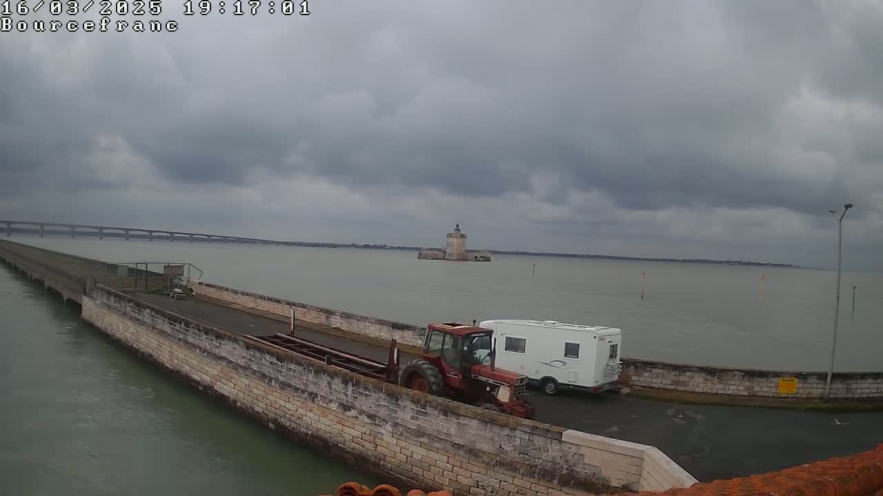 A tractor and campervan are parked on a stone causeway extending into a body of water under a cloudy sky, with a distant bridge and a small tower visible.