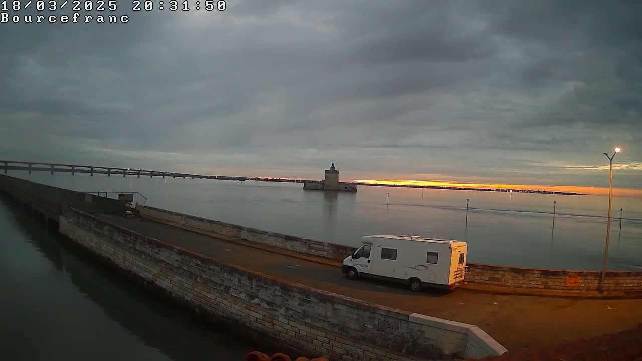 A white camper van is parked on a stone wall overlooking calm water at sunset, with a distant bridge and stone tower visible.