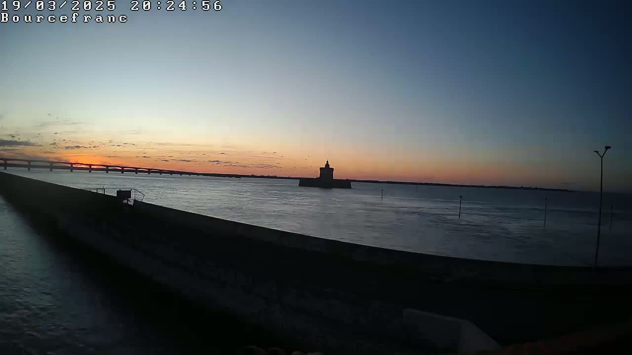 A long, dark wall extends into a calm body of water at sunset, with a distant bridge and a silhouetted tower visible on the horizon.