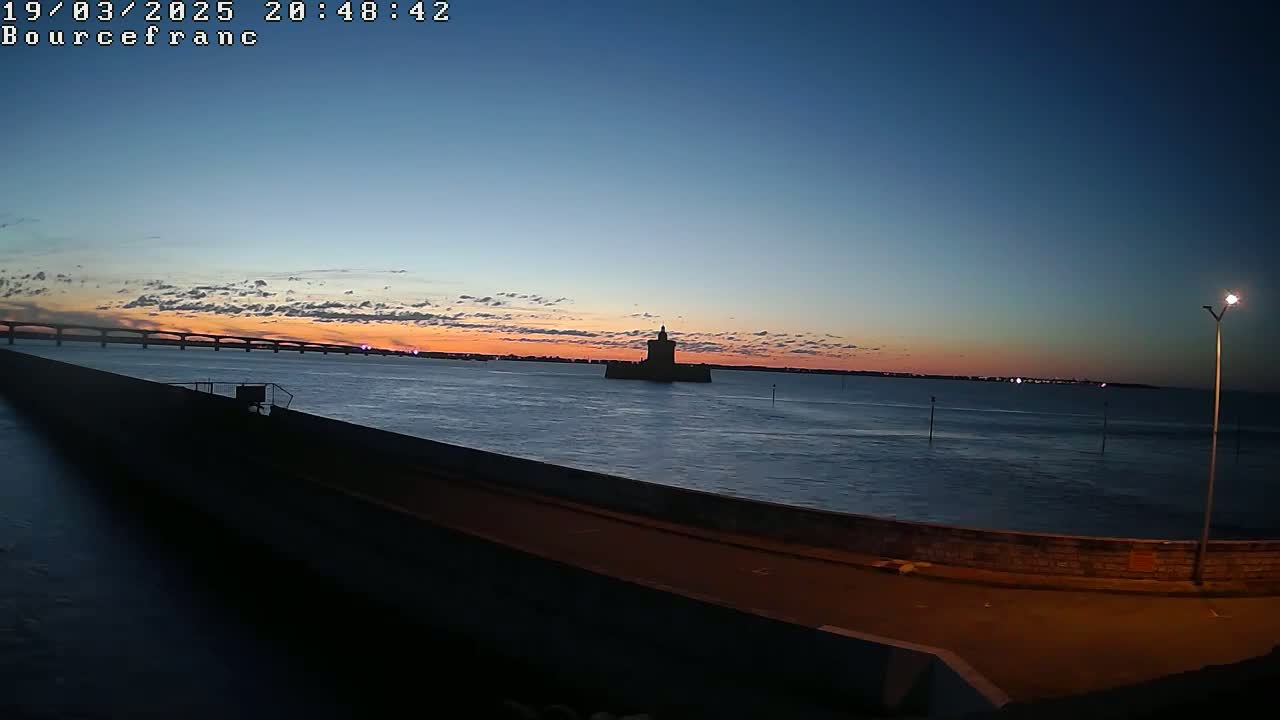 A dark silhouette of a tower stands in the water at sunset, with a bridge visible in the distance under a mostly clear sky.