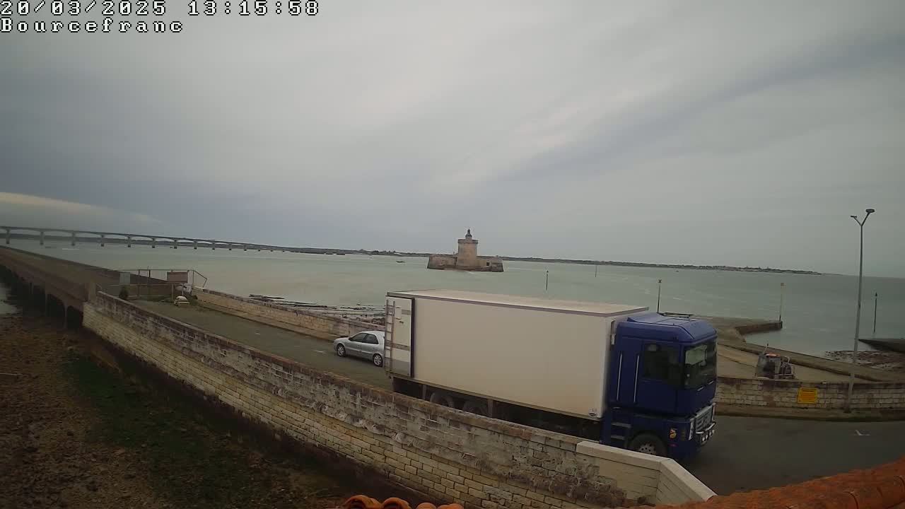 A blue truck and a car are parked near a stone wall overlooking a body of water with a distant bridge and a stone tower under an overcast sky.