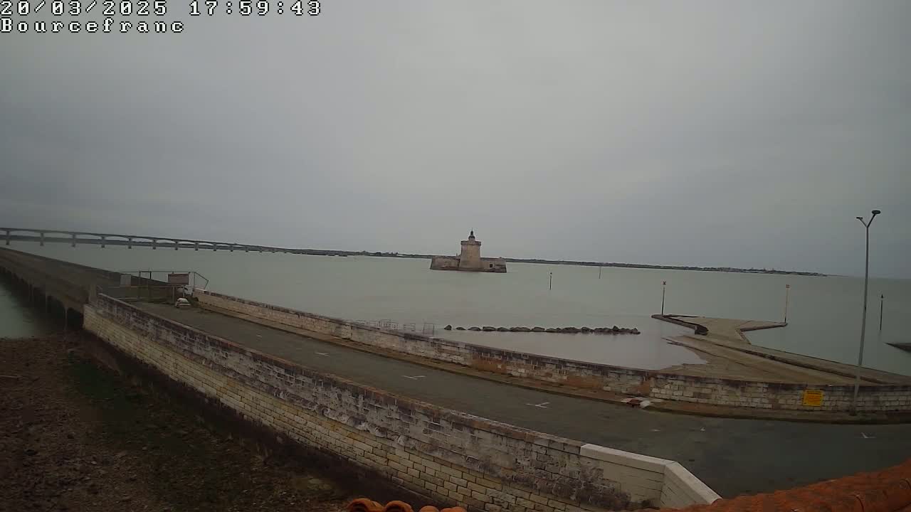 A stone pier extends into a calm, gray-sky day over a body of water, toward a small stone tower, with a bridge visible in the far background.