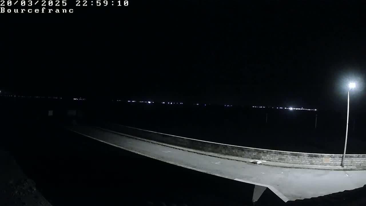 A nighttime view shows a long, light-colored road or path extending into the distance, bordered by a low wall, with distant lights faintly visible on the horizon under a dark sky.