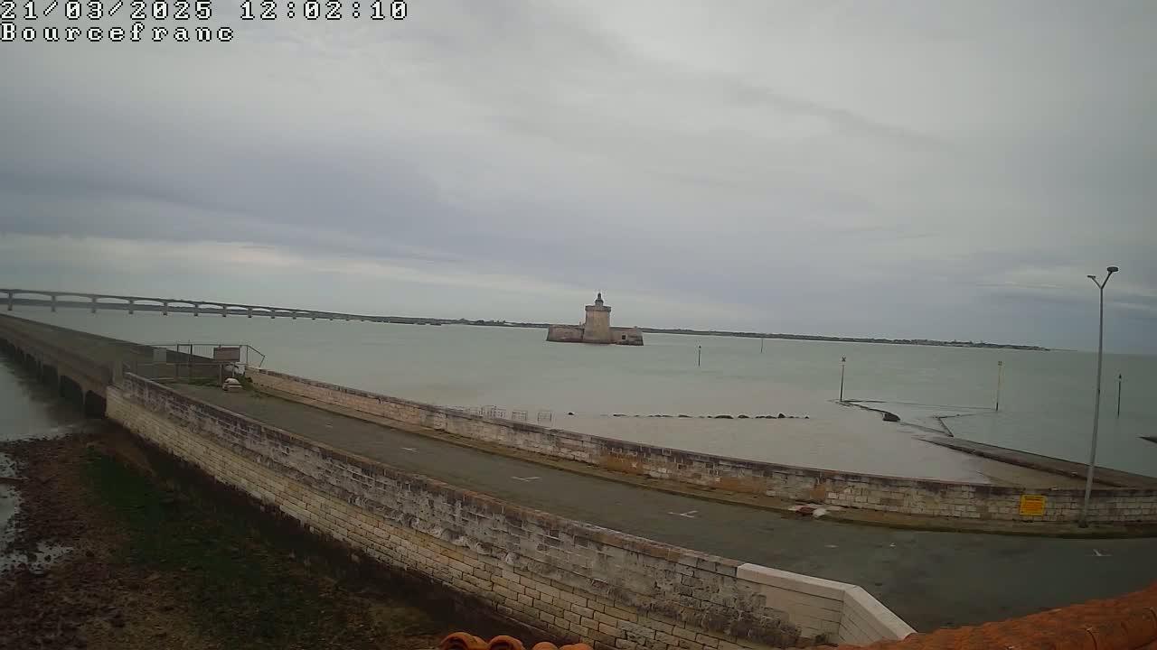 A stone wall and roadway extend into a calm, gray-sky body of water, toward a distant stone tower and a long bridge.