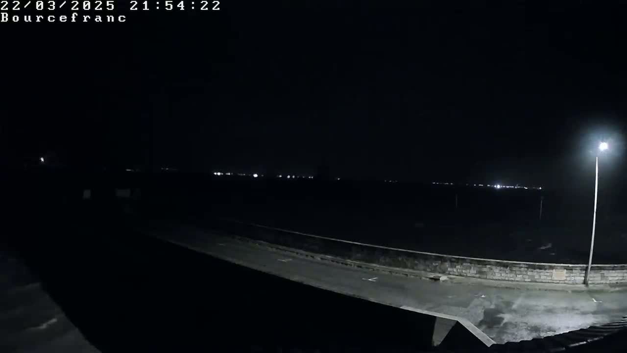 A nighttime, low-light view shows a paved area, a low wall, and a distant horizon with faint lights under a dark sky.