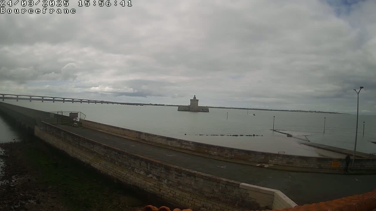 A stone wall extends into a body of water under a cloudy sky, with a distant bridge and a stone tower visible in the background.