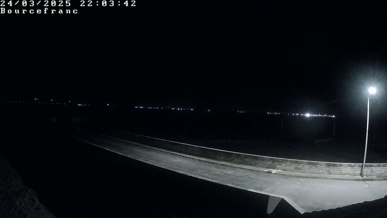 A nighttime view of a road and low wall illuminated by a single lamppost, with distant lights visible on the horizon.