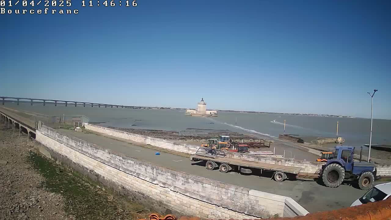 A long bridge stretches across a calm, light-grey body of water under a clear blue sky, with several tractors and a flatbed trailer parked near a stone wall in the foreground, and a small tower visible in the mid-ground.