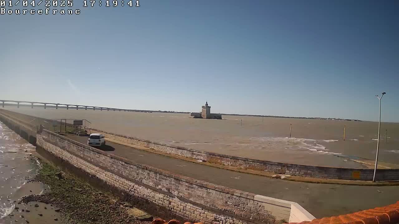 A distant stone tower sits on a calm, light-brown body of water under a clear blue sky, viewed from a stone wall next to a road, with a bridge visible in the background.