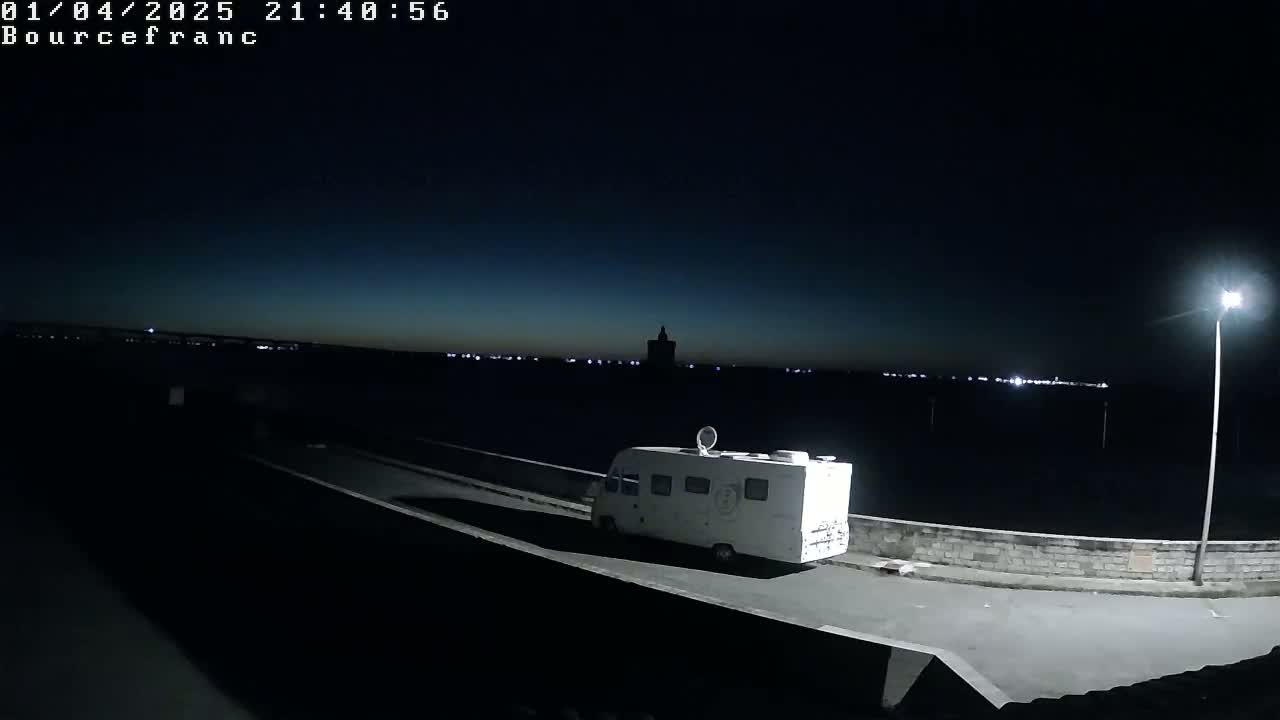 A white van is parked on a dark road at night next to a low wall, with distant lights visible on the horizon under a mostly clear, dark sky.