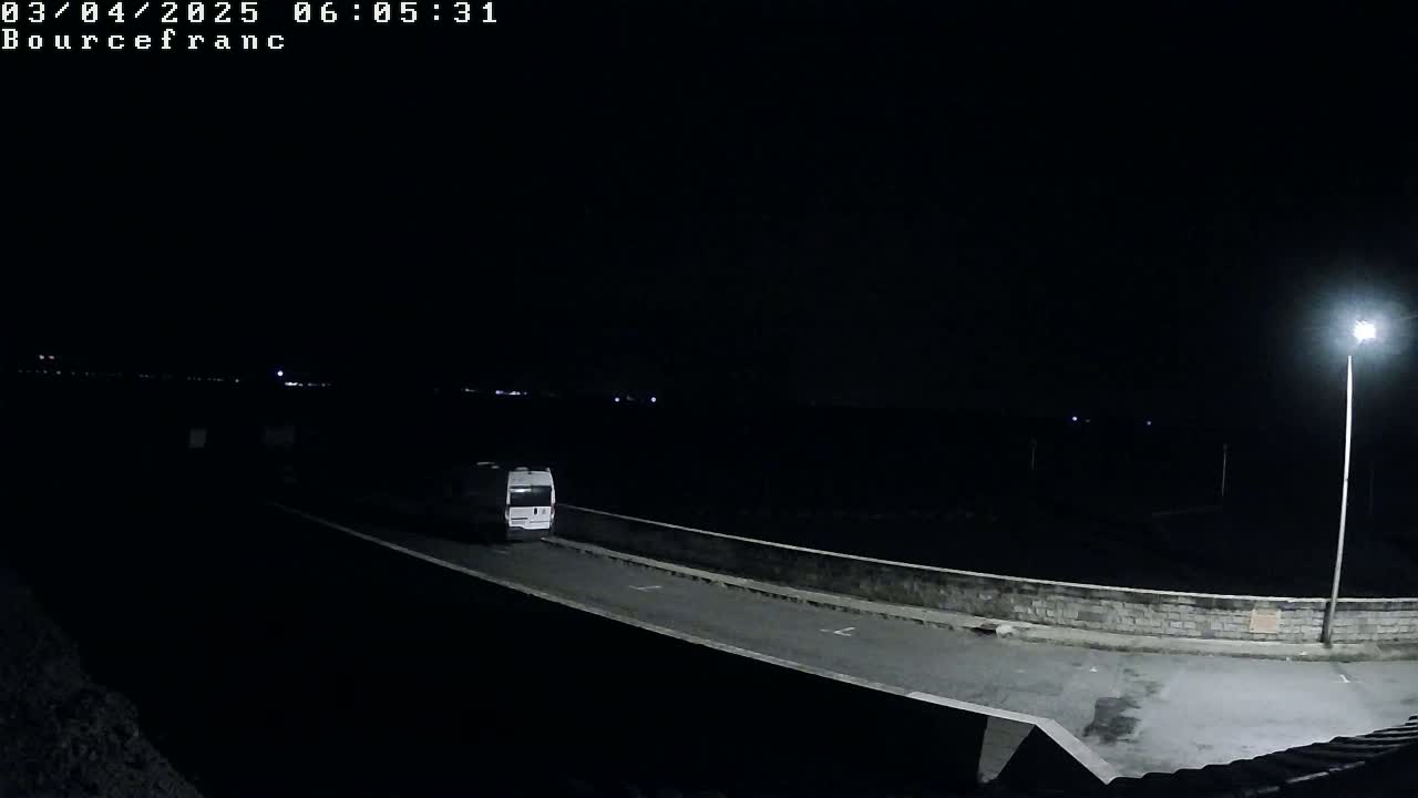 A white van drives on a dark road at night illuminated by a single streetlight.