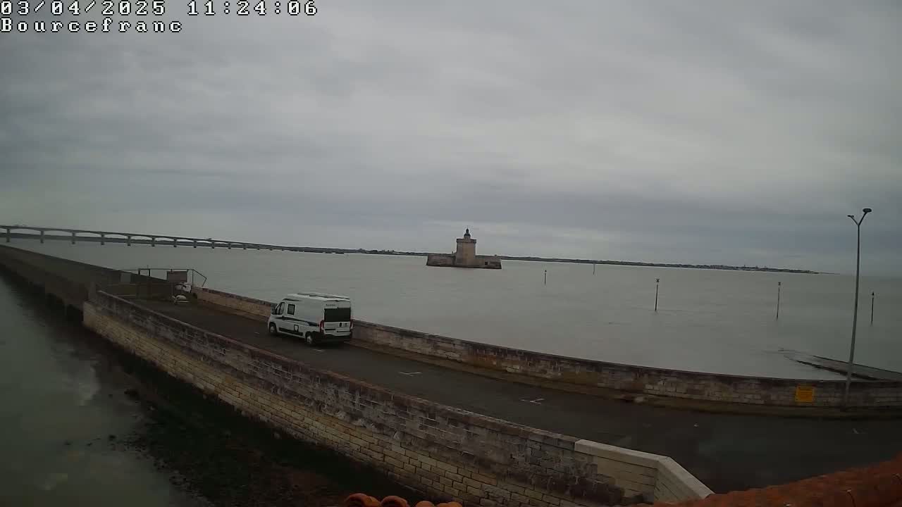 A white camper van is parked on a stone wall beside a calm body of water under an overcast sky, with a distant bridge and a stone tower visible in the background.