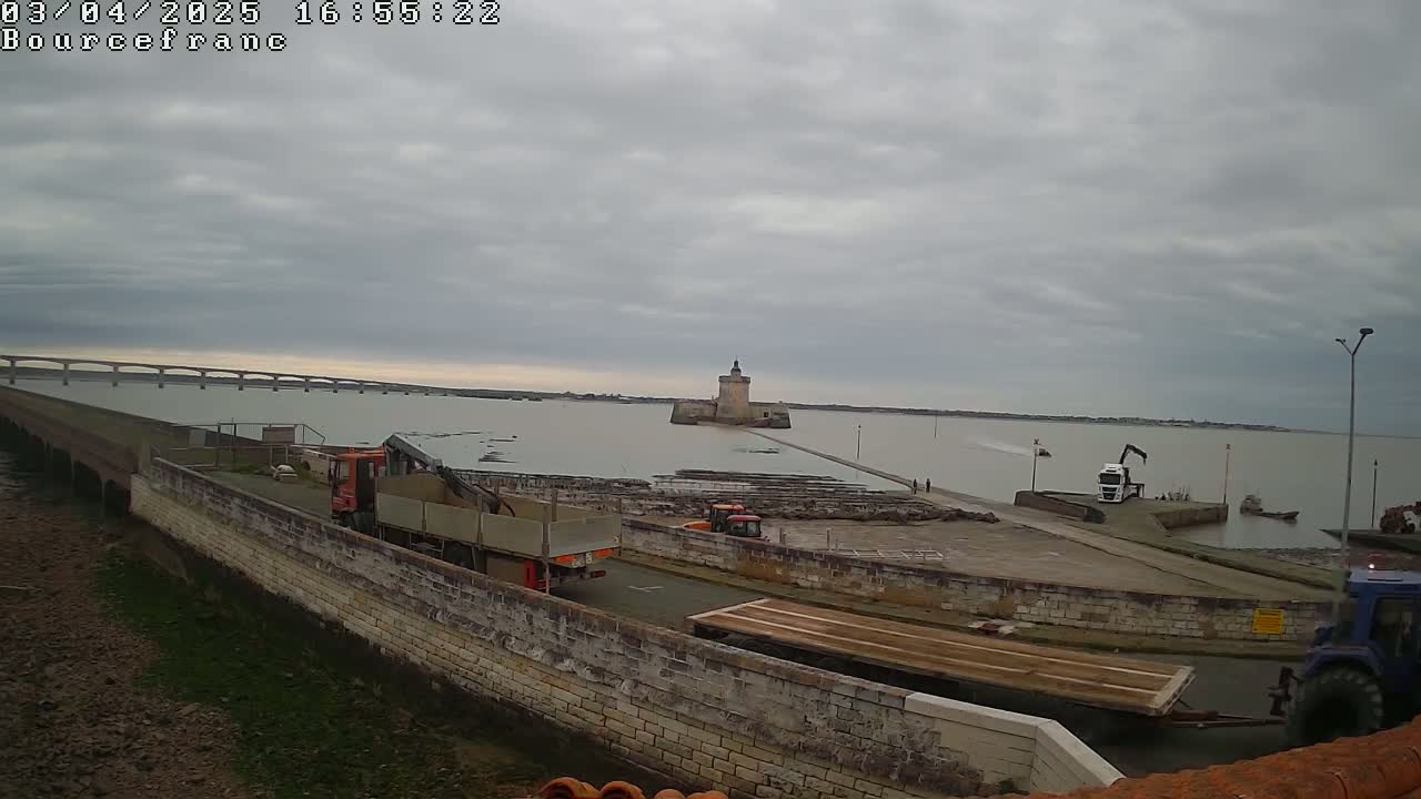 A stone tower sits on a body of water under an overcast sky, with trucks and other construction equipment on the shore and a long bridge visible in the distance.