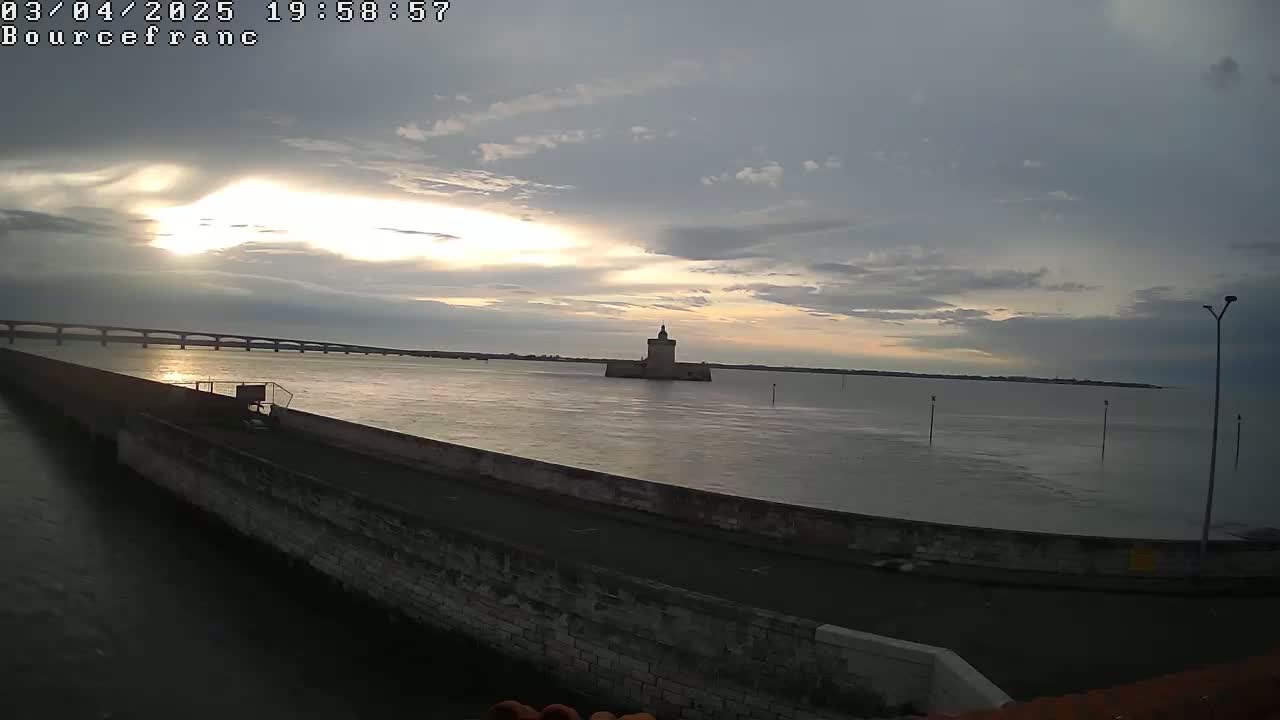 A stone tower sits in calm water under a partly cloudy sky at sunset, with a long bridge visible in the distance.