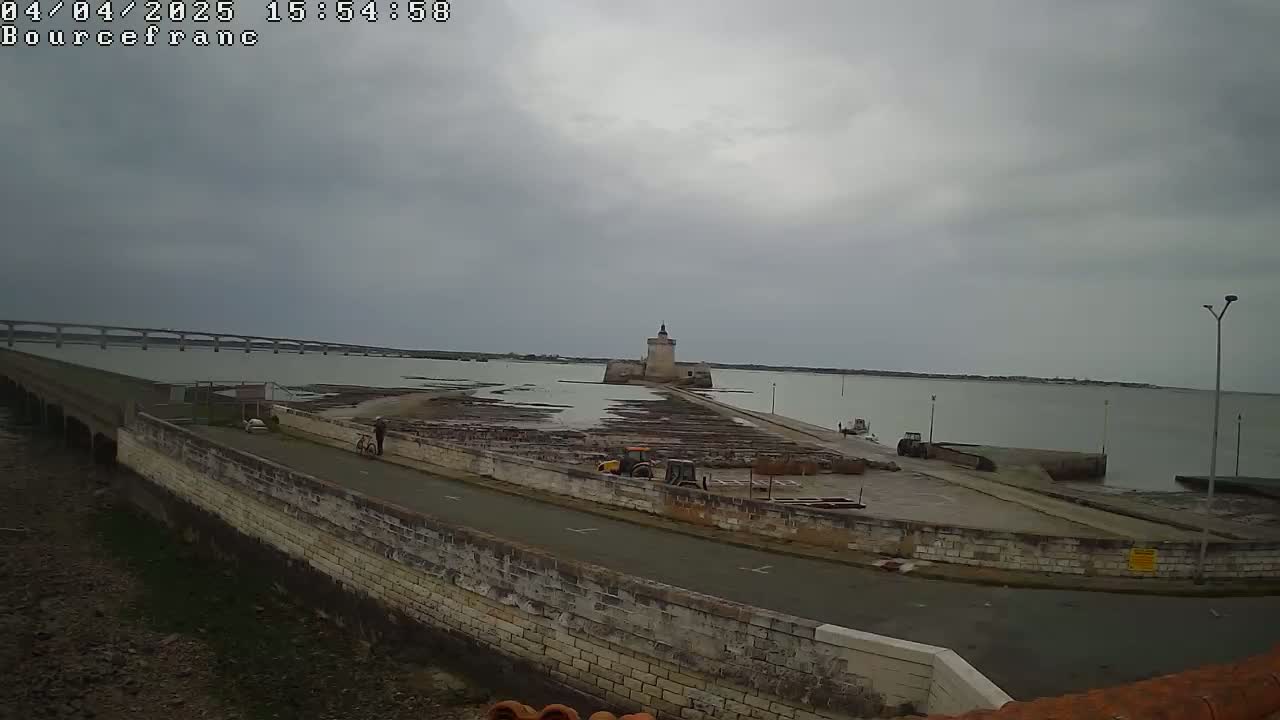 A stone tower sits on a causeway extending into a calm, gray-sky day over a body of water, with a bridge visible in the distance and some small vehicles near the tower.