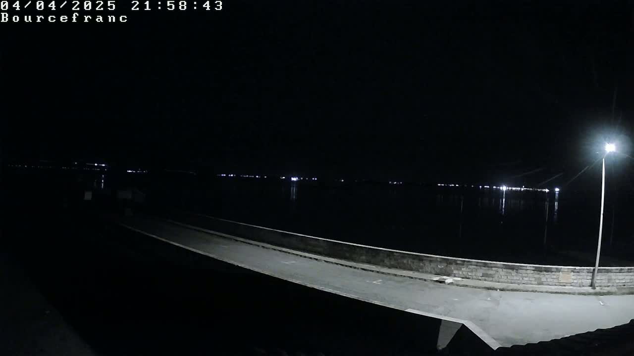 A nighttime view of a paved walkway extending over dark water, illuminated by a single lamppost and distant lights reflecting on the water.