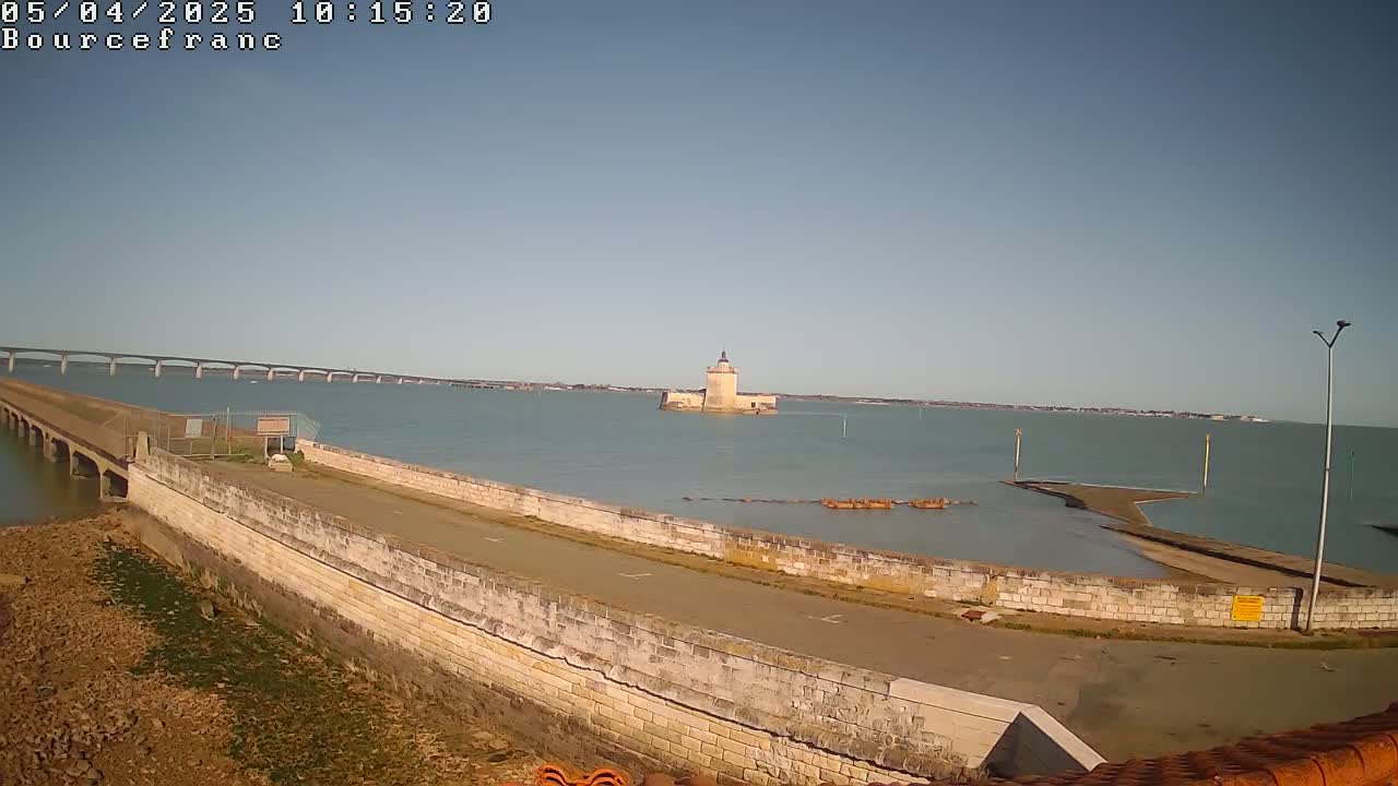 A stone seawall extends into a calm body of water, featuring a distant tower and a long bridge under a clear, sunny sky.