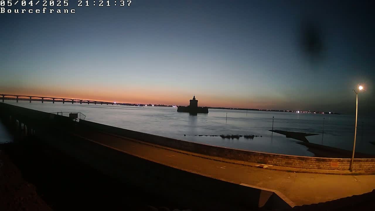 A dark silhouette of a tower stands in the middle of a calm body of water during twilight, with a bridge visible in the distance under a pinkish-orange sky.