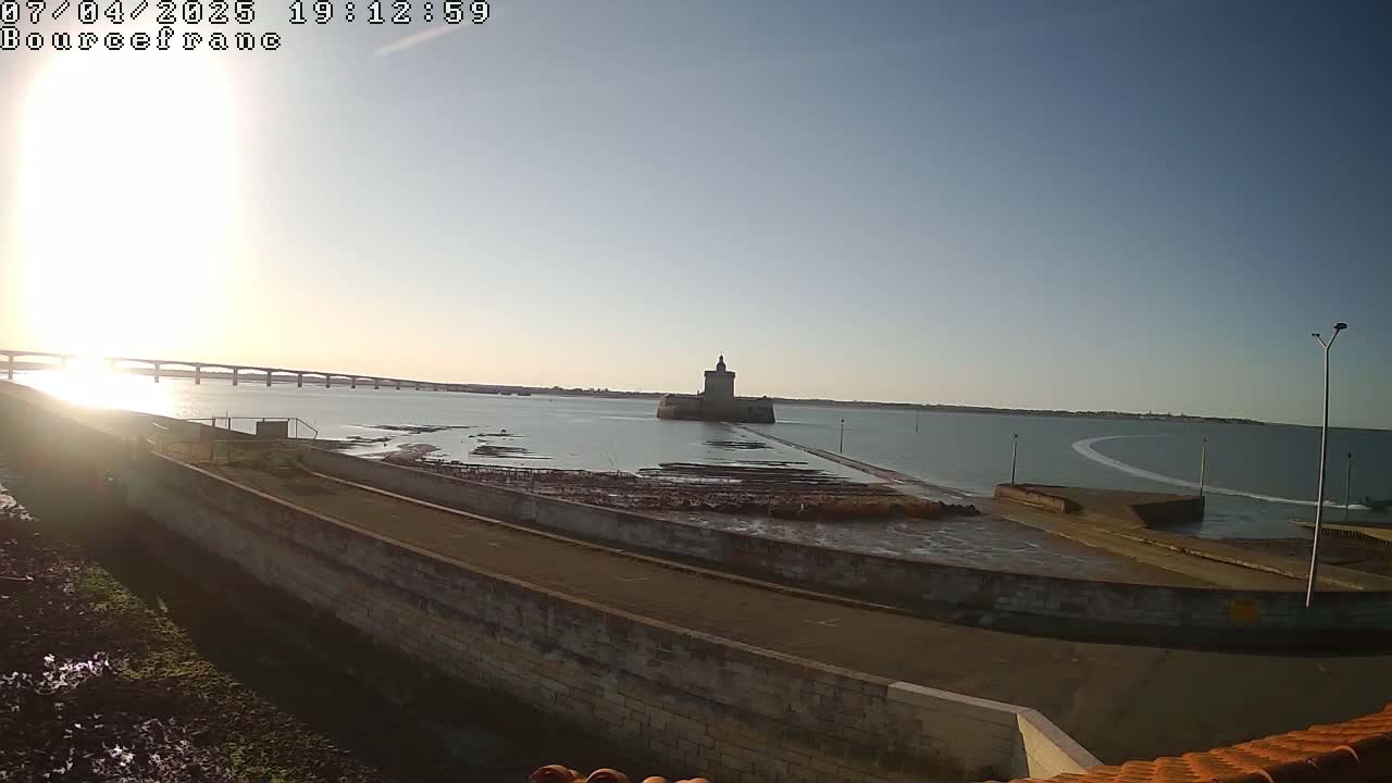 A stone fortress sits in calm water at sunset, with a bridge visible in the distance and low tide revealing exposed seabed.