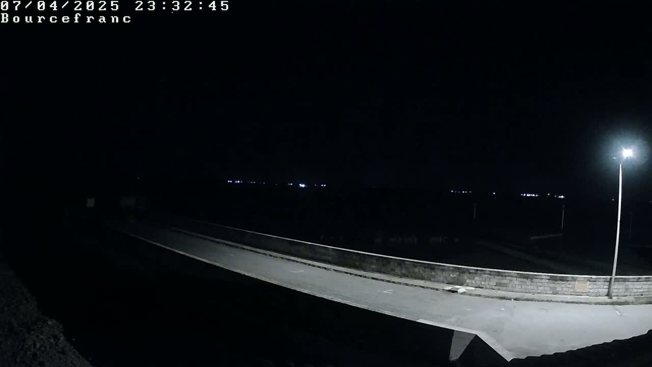 A nighttime, low-angle view shows a paved road curving gently around a low stone wall, with distant lights visible in the darkness.