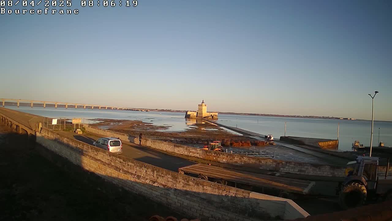 A long bridge spans a calm body of water under a clear, bright sky, with a stone tower and causeway in the foreground, and a tractor and other vehicles near a low stone wall.
