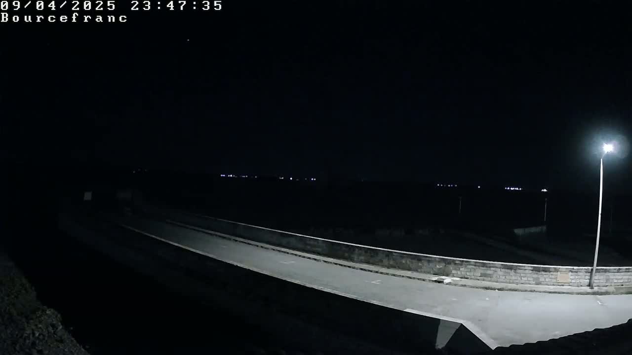 A nighttime view of a lightly-lit road curving around a low wall, with a distant horizon showing faint lights under a dark sky.