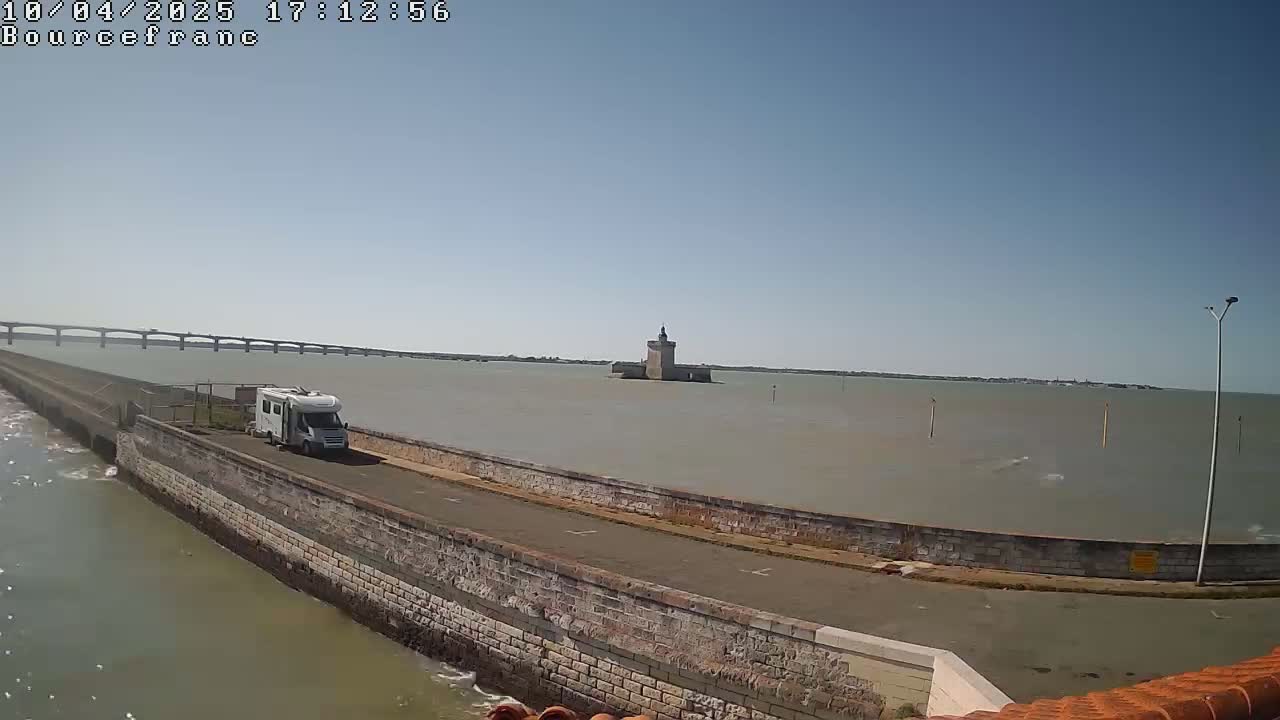 A white camper van is parked on a stone roadway next to a body of water, with a distant bridge and a stone tower visible under a clear sky.