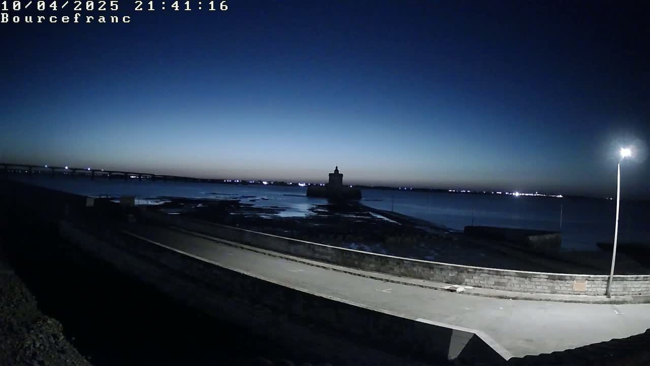 A dark, low-light view shows a stone roadway curving alongside a body of water, with a distant bridge and a dark tower in the background under a twilight sky.