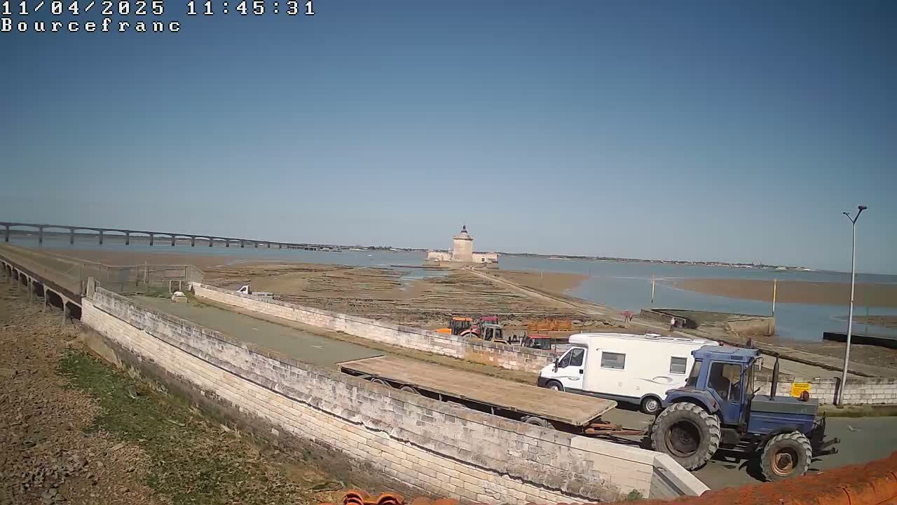 A coastal scene on a sunny day shows a stone causeway leading to a tower, construction equipment, and a camper van, with a bridge visible in the distance.