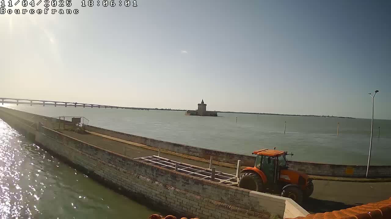 An orange tractor is parked near a stone wall overlooking calm water, a distant bridge, and a stone tower on an island.