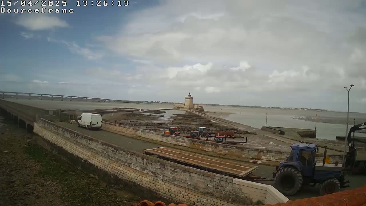 A stone causeway leads to a stone tower on an island, with a bridge visible in the distance under a mostly cloudy sky; several tractors and a van are present.