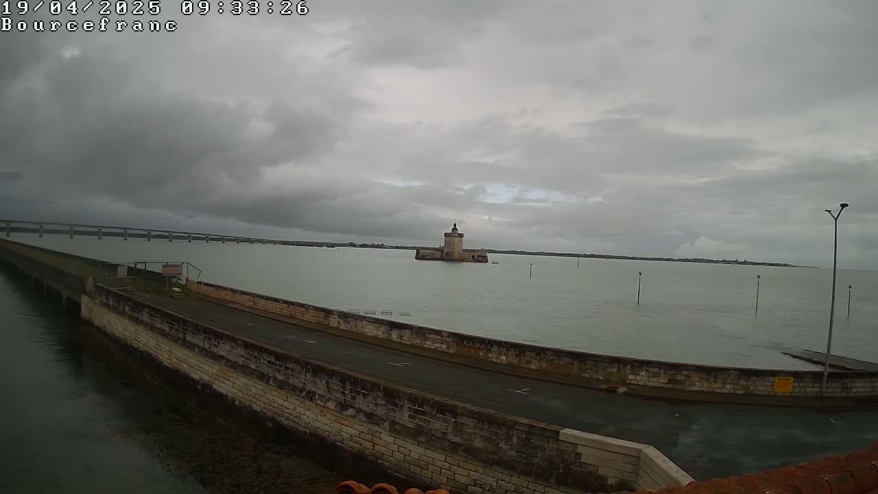 A stone tower sits in a gray, overcast body of water, with a long bridge visible in the distance.