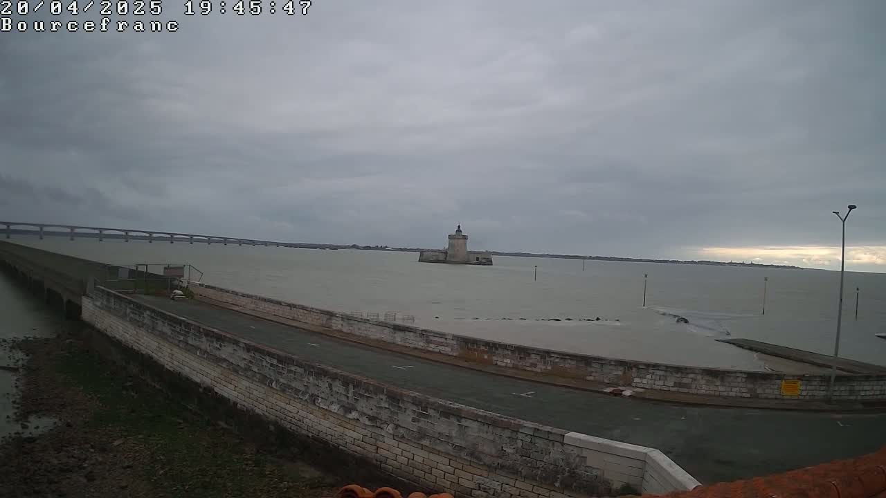 A stone tower sits in a calm body of water under an overcast sky, with a long bridge visible in the distance.