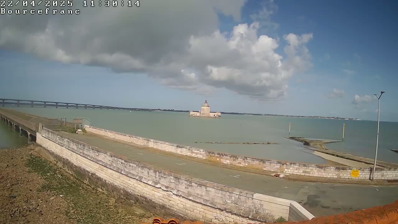 A stone causeway leads to a small island with a stone tower in a calm body of water under a partly cloudy sky, with a bridge visible in the background.
