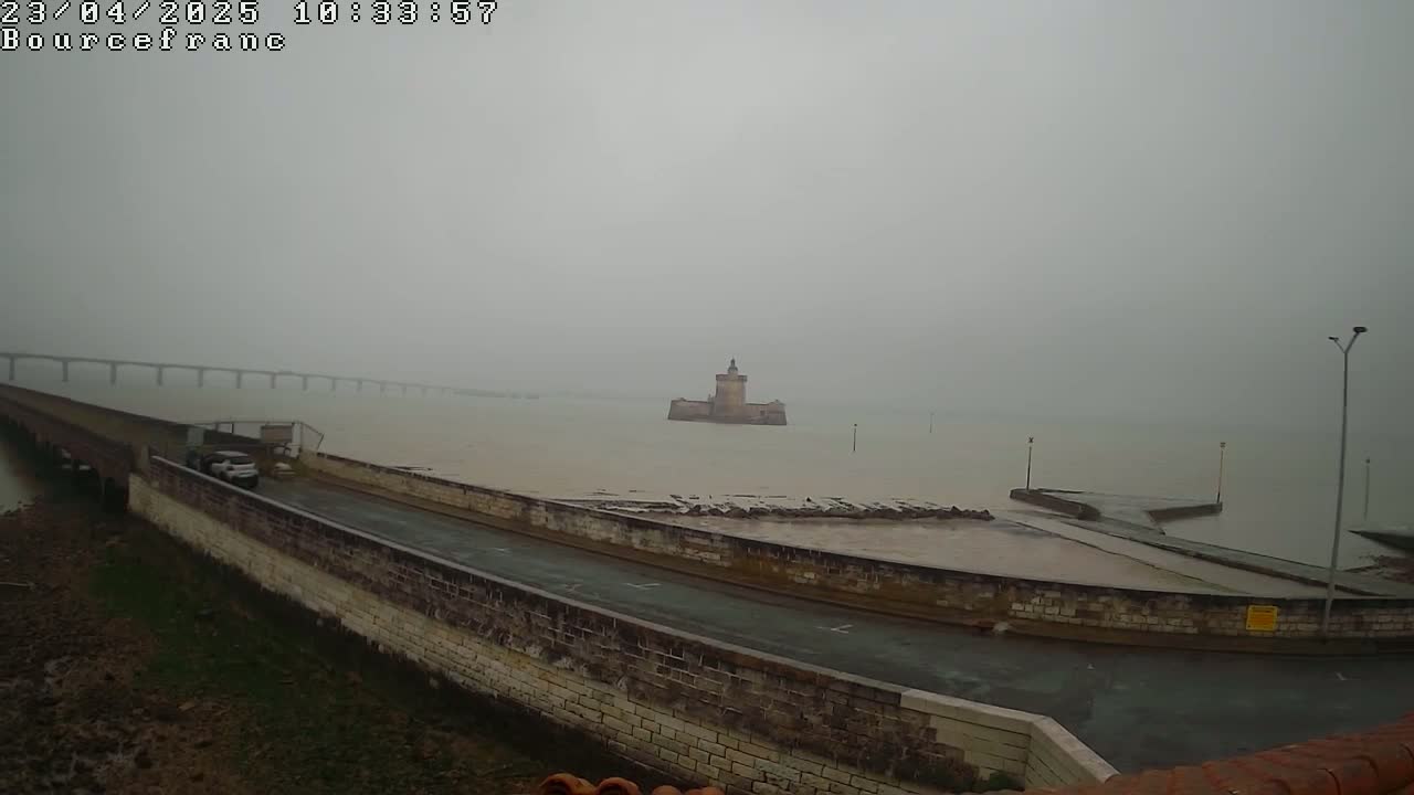 A stone wall and road overlook a calm, foggy body of water, a distant bridge, and a small stone fortress.