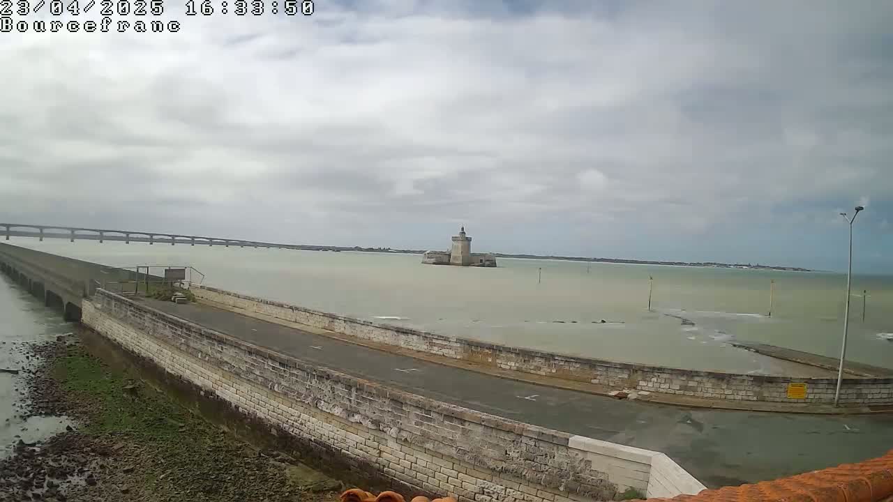 A stone seawall curves around a roadway, overlooking a calm body of water where a stone tower sits in the distance, under an overcast sky.