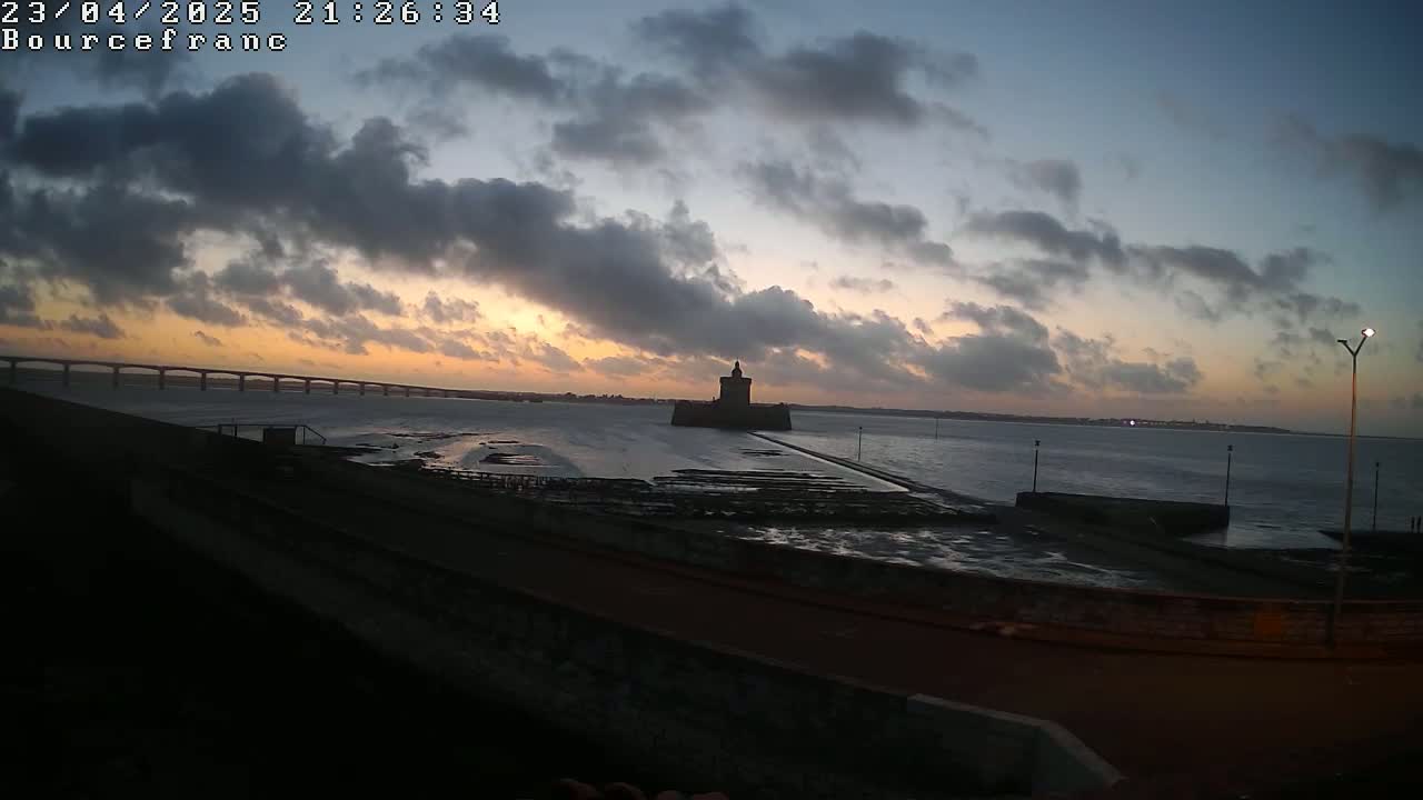 A dark silhouette of a stone tower is visible on the coast, under a partly cloudy sunset sky, near a bridge and calm water.