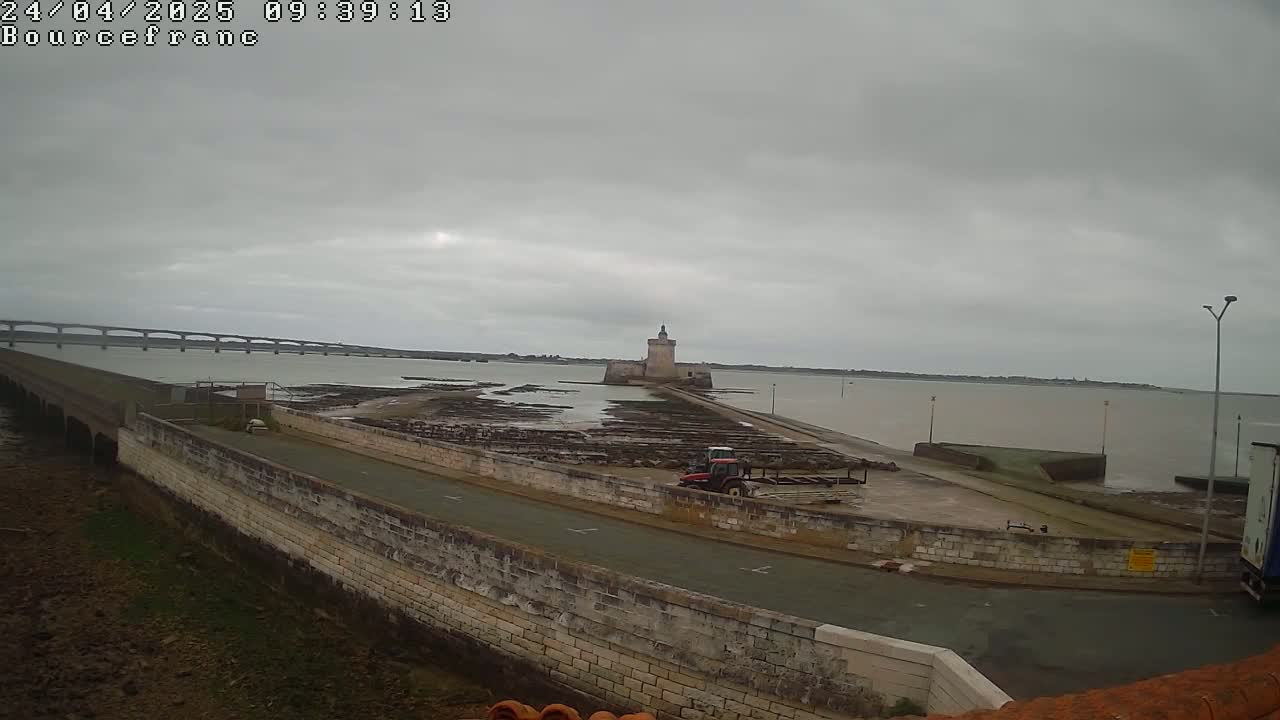 A stone tower sits on a causeway extending into calm, gray water under an overcast sky, with a bridge visible in the distance.