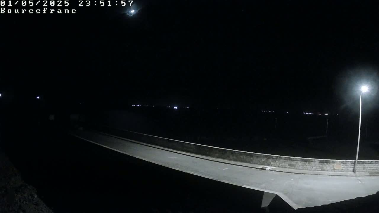 A nighttime view shows a long, empty road or bridge illuminated by a single streetlight, with faintly visible lights in the distance under a dark sky.