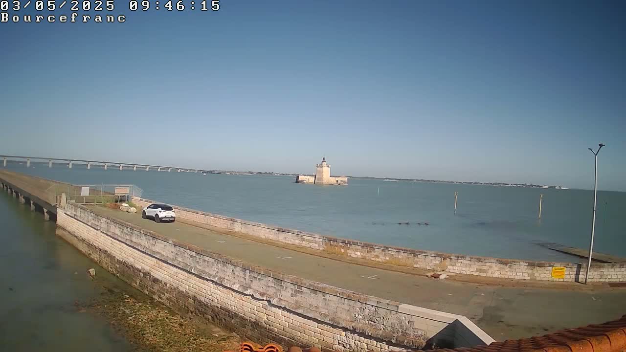 A stone seawall curves toward a distant tower on a calm, sunny day, with a bridge visible in the background and a single car parked nearby.