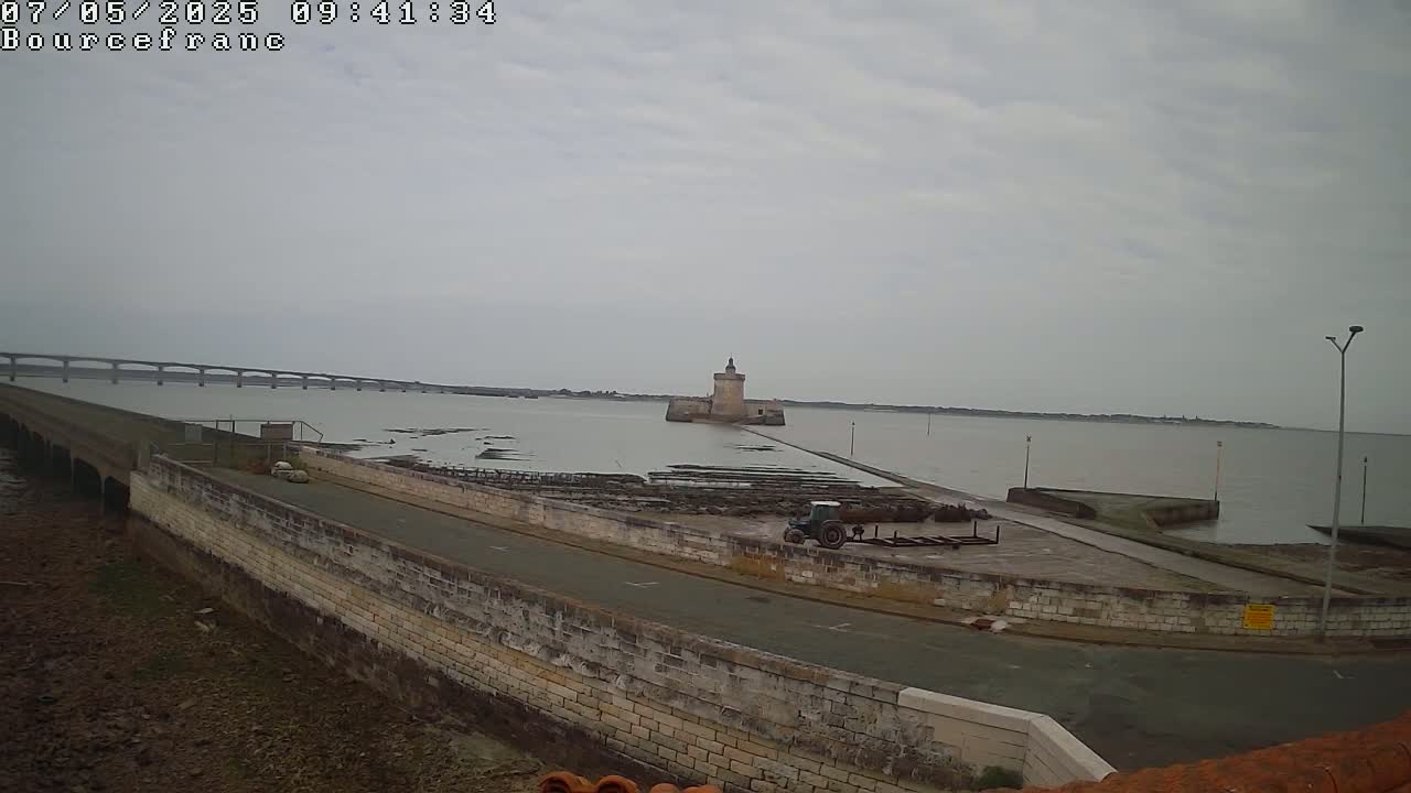 A stone tower sits in calm, overcast waters, connected to a long causeway leading to a distant bridge.