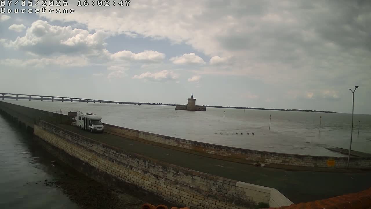 A motorhome is parked on a stone wall overlooking calm water, a distant bridge, and a small stone tower under a partly cloudy sky.