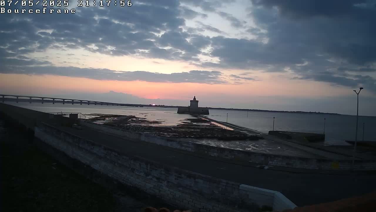 A stone tower sits on a causeway extending into a calm body of water at sunset under a partly cloudy sky, with a bridge visible in the distance.