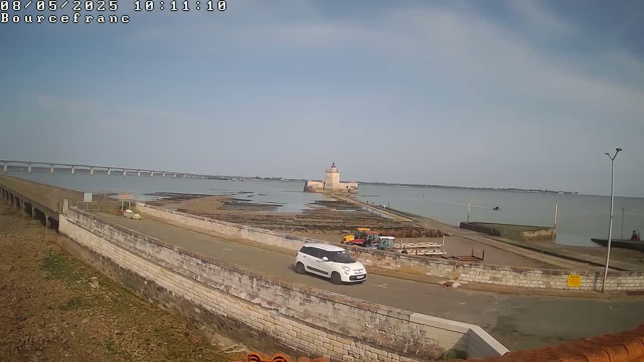 A white car drives on a road leading to a stone fort on a calm, sunny day, with a long bridge visible in the distance and farm equipment nearby.