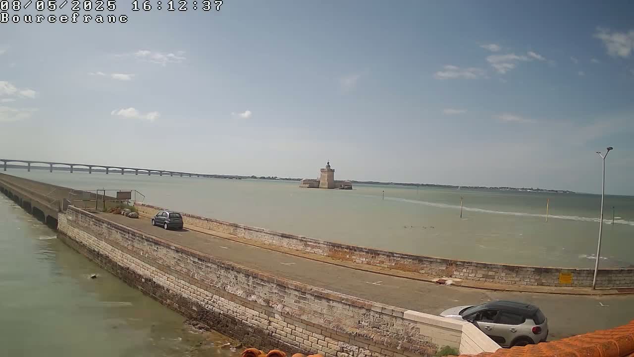 A stone wall and road border a body of water, with a distant bridge and a small, stone tower visible under a partly cloudy sky.
