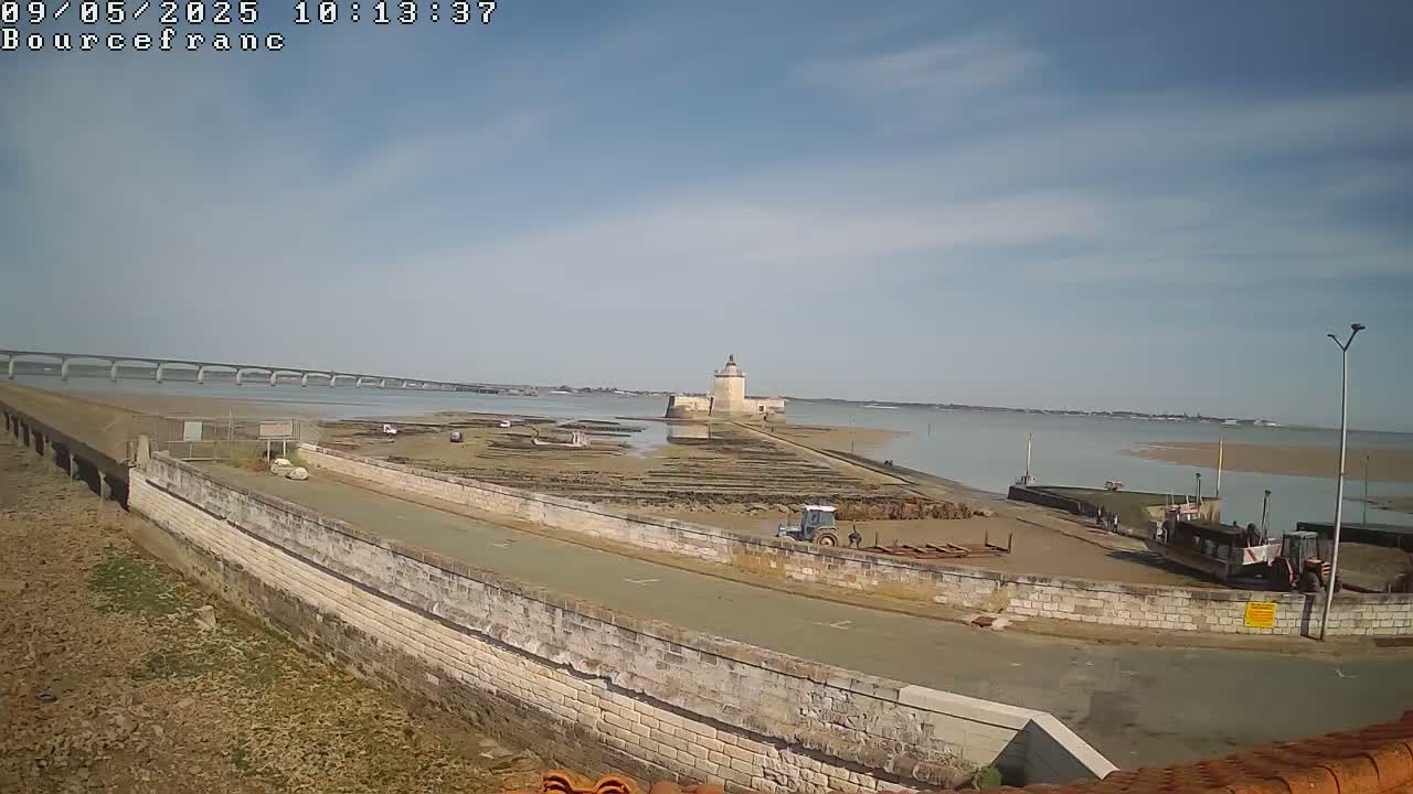 A stone wall curves around a harbor area with a tractor, a distant fort, and a long bridge visible under a mostly sunny sky.