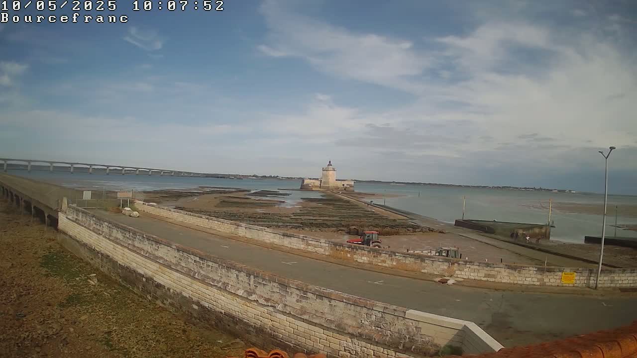 A stone wall and road lead to a tower on an island connected to the mainland by a causeway, with a bridge visible in the distance under a partly cloudy sky.