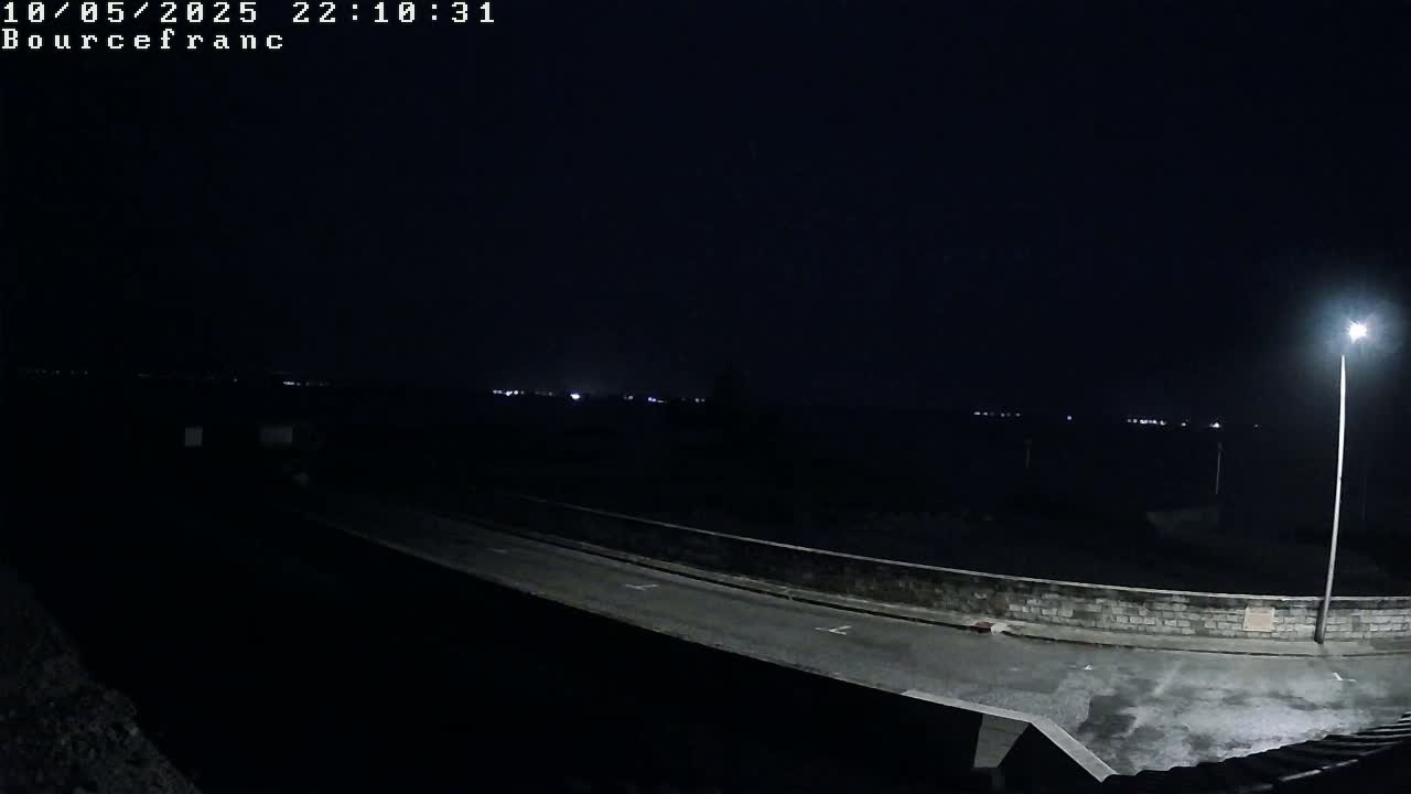 A nighttime view of a dark road illuminated by a single lamppost, overlooking a low wall and a distant line of faint lights.