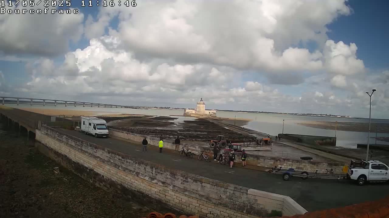 A coastal scene under a partly cloudy sky shows a stone structure with a tower in the middle of a body of water, a bridge in the distance, several people and vehicles near the shore, and a campervan parked on a stone path.